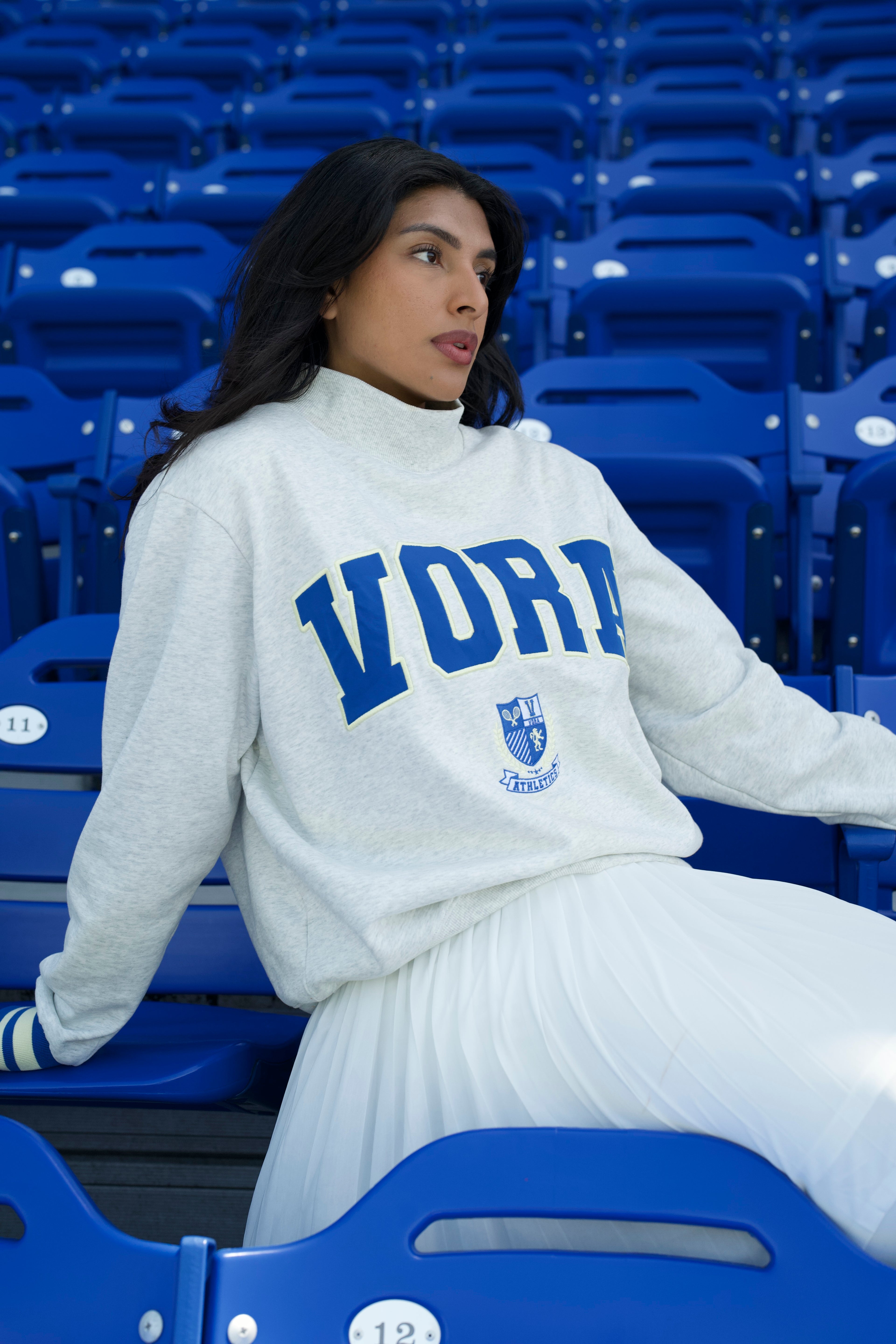 Woman wearing a white sweatshirt with 'Vora' printed on it, sitting in a stadium with blue seats.